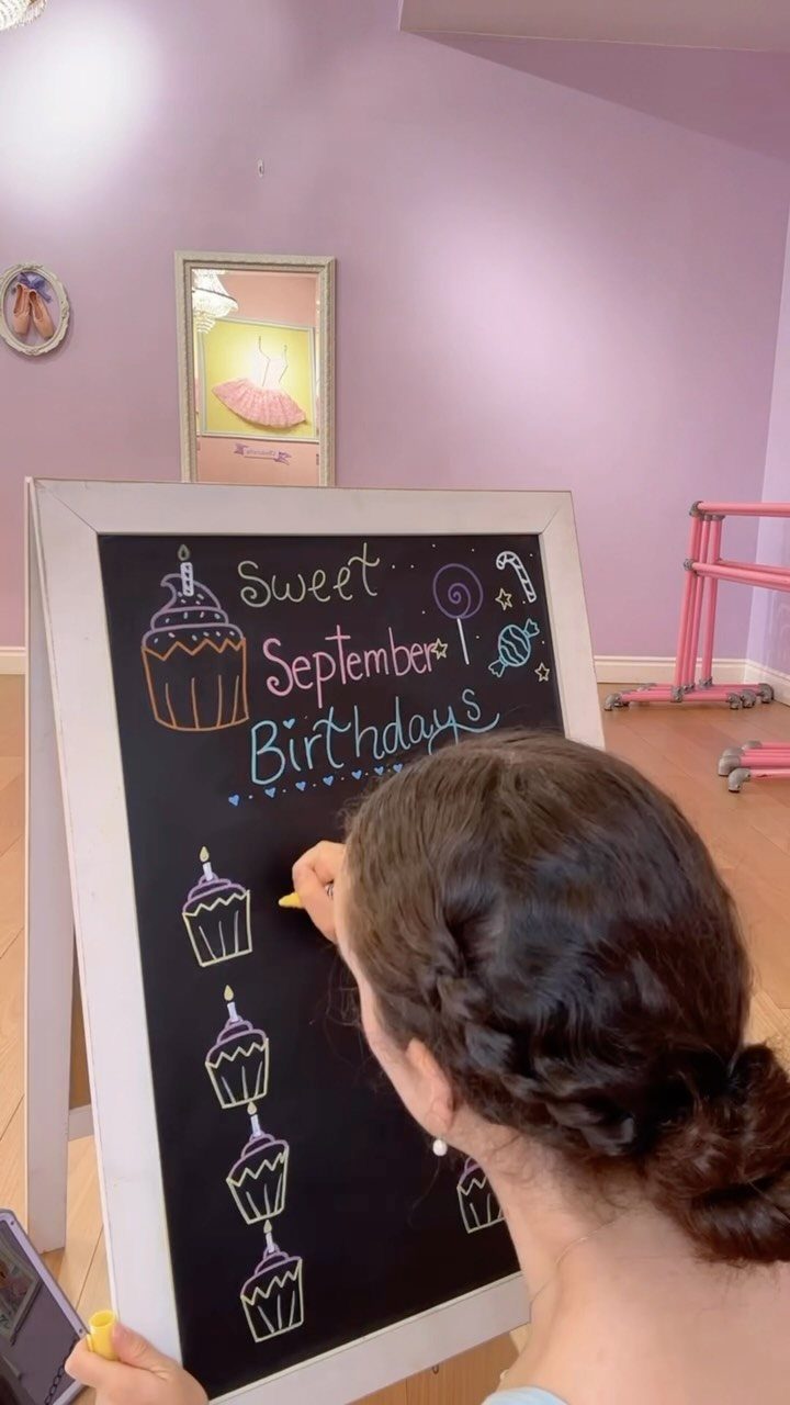 Woman drawing colorful cupcakes and birthday messages on a white-framed chalkboard sign.