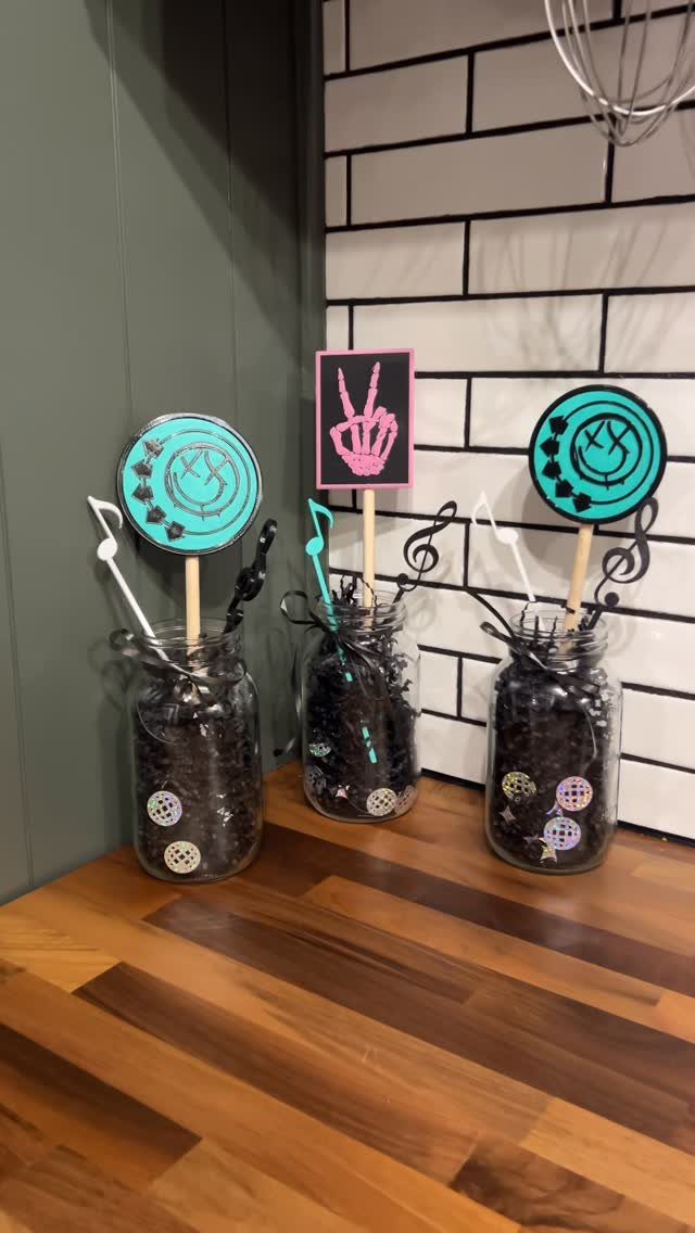 Three mason jar centerpieces with black filler and themed toppers on a wooden floor.