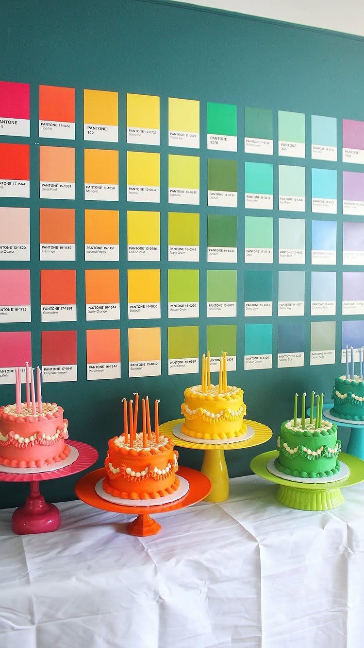 Colorful birthday cakes on pedestals in front of a rainbow paint swatch wall.