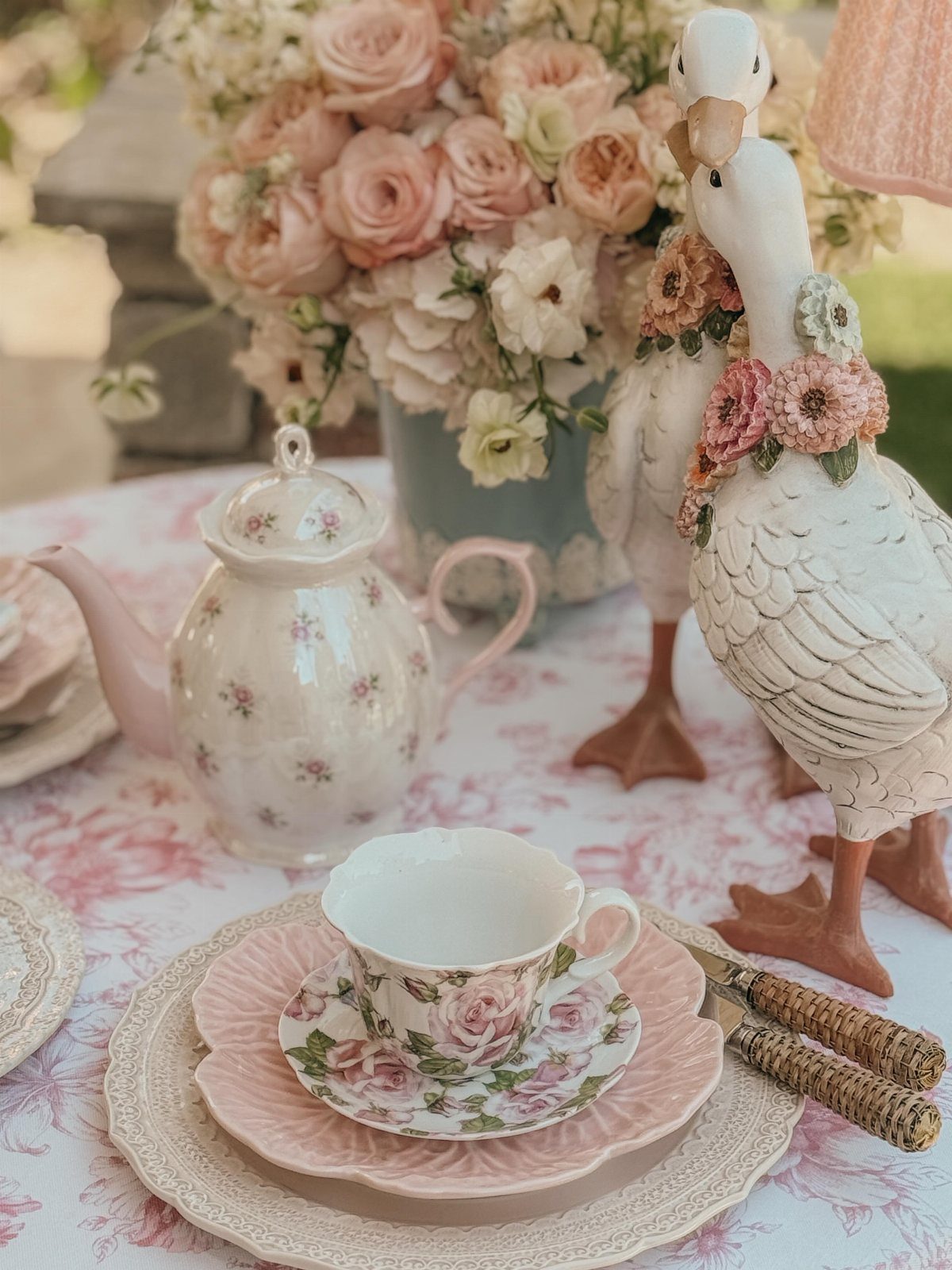 Floral tea party table setting with pink roses and white goose figurines.