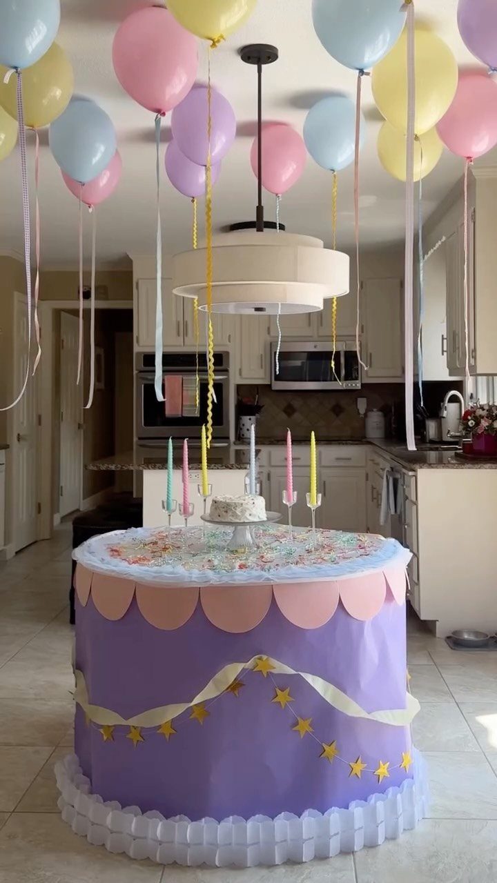 Purple cake-shaped party table with pastel balloons and candles in a kitchen setting.