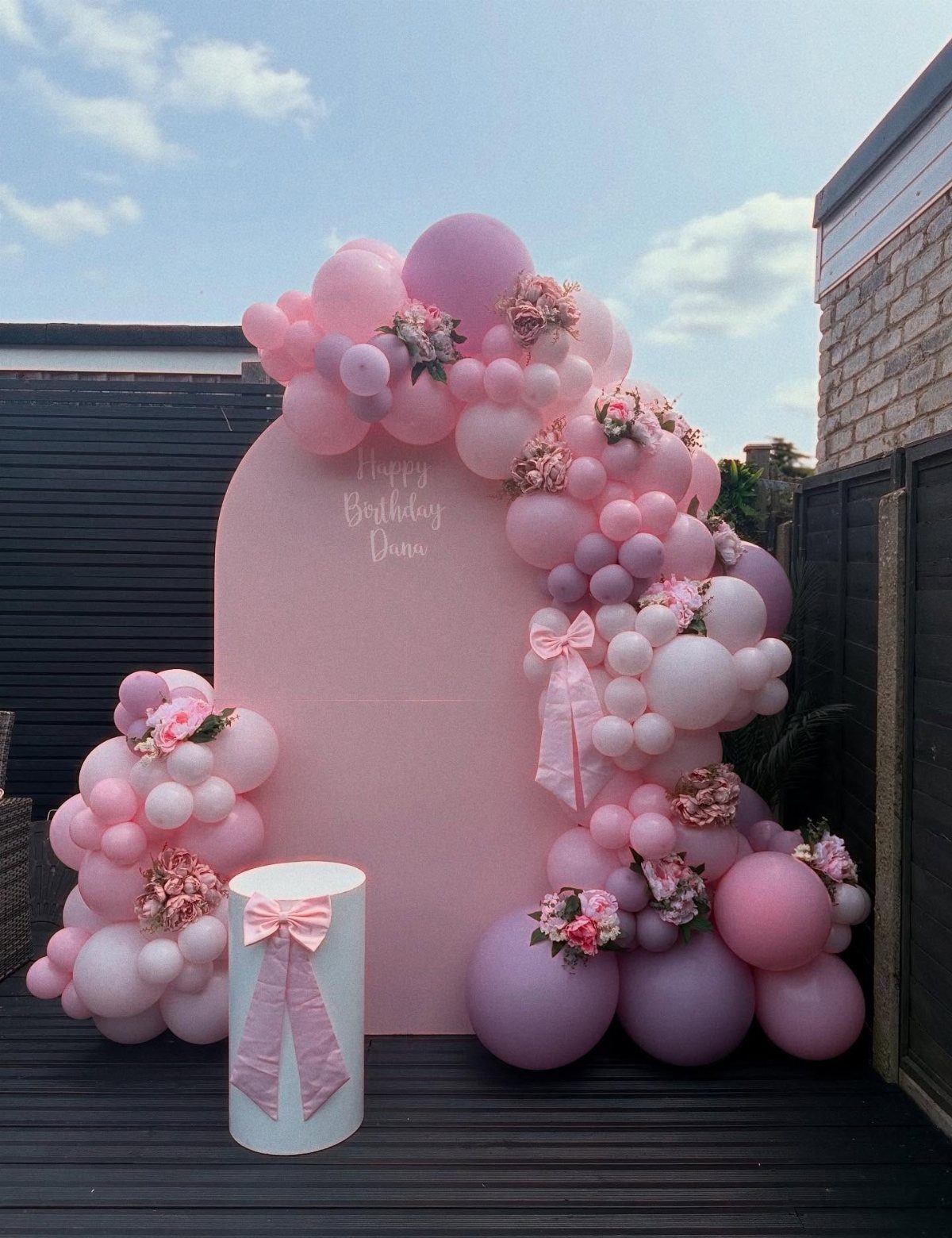 Pink arched backdrop with a colorful balloon garland and floral accents outdoors.