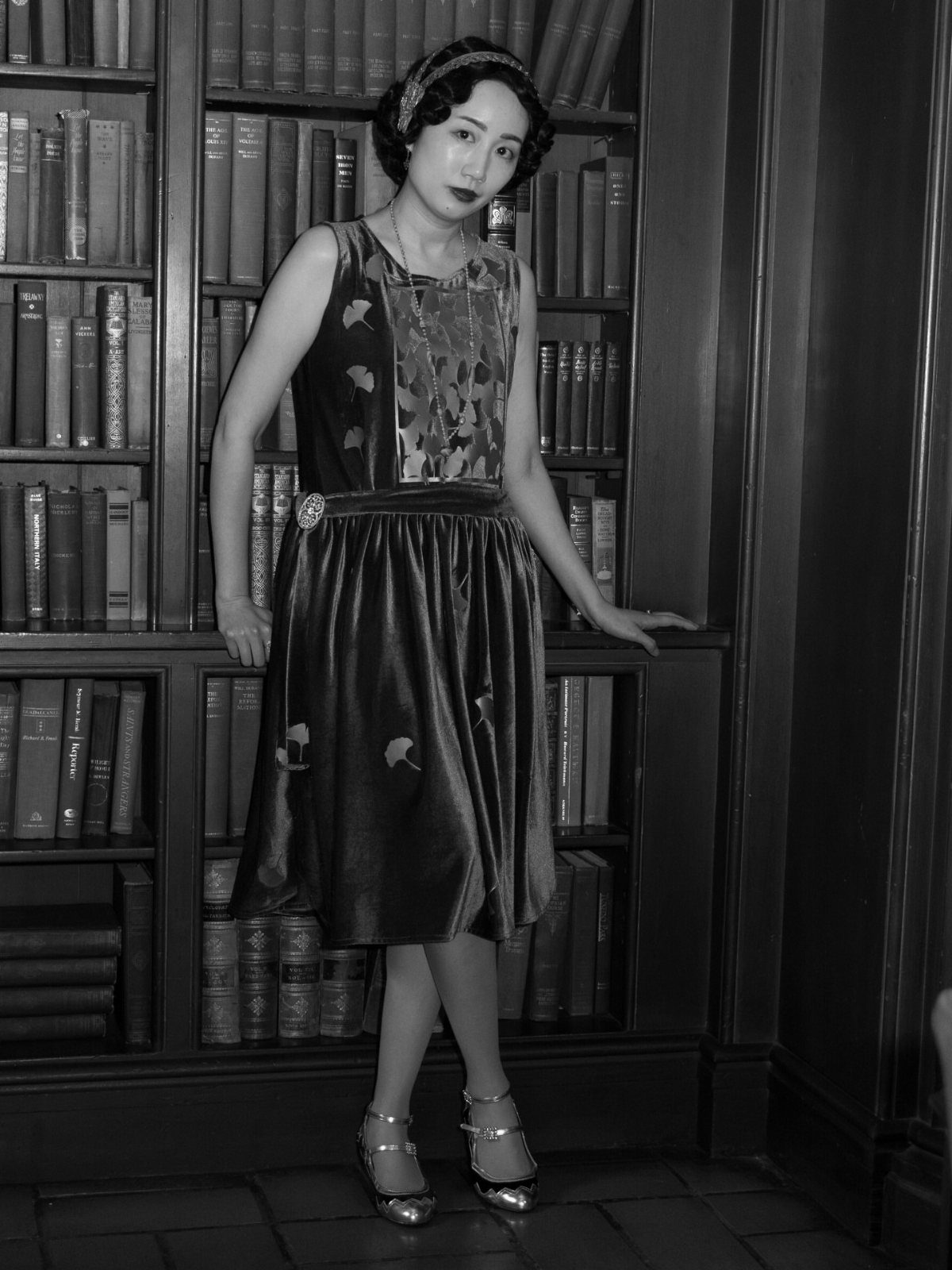 Woman in vintage 1940s style dress and headband posing by a bookshelf.