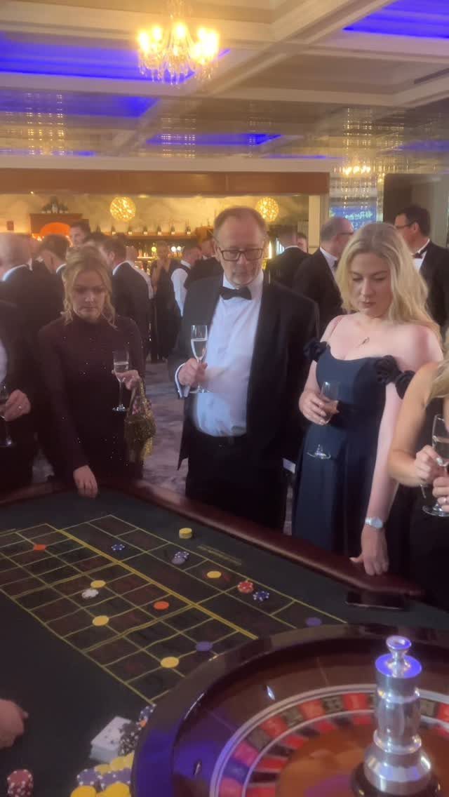 People in formal black and white attire standing around a casino roulette table.