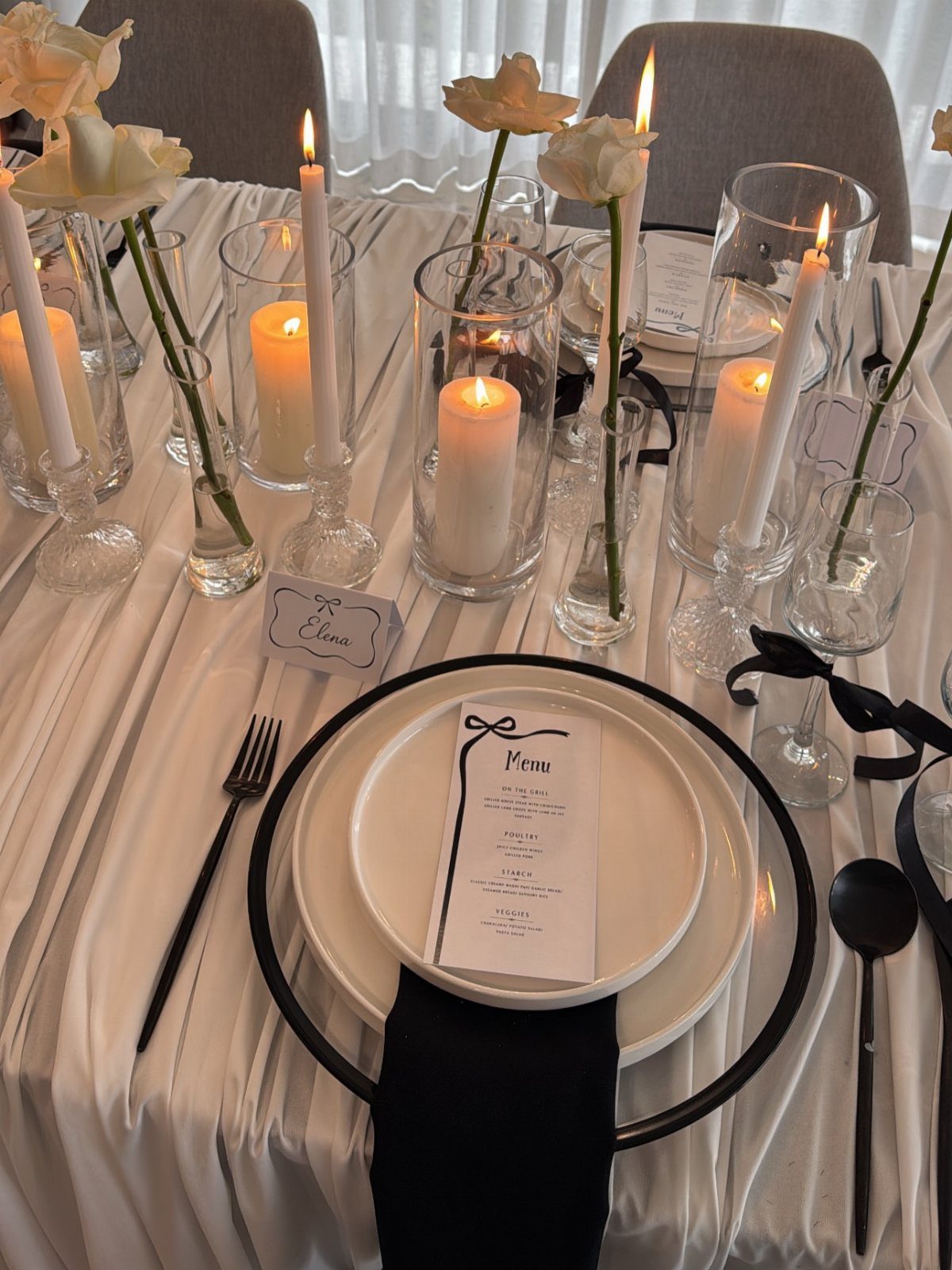 Formal table setting with white plates, black cutlery, and white candles and flowers.