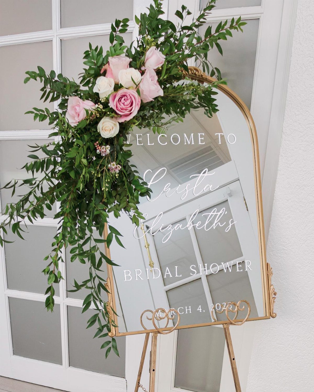 Arched gold mirror welcome sign for a bridal shower decorated with pink roses.