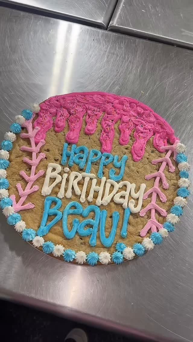 Cookie cake with pink frosting drips and blue birthday lettering on metal surface