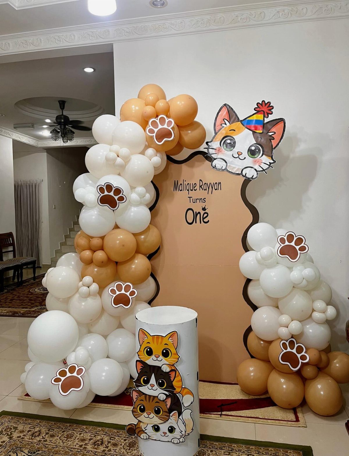 Birthday backdrop with tan and white balloon arches and cartoon cat decorations.