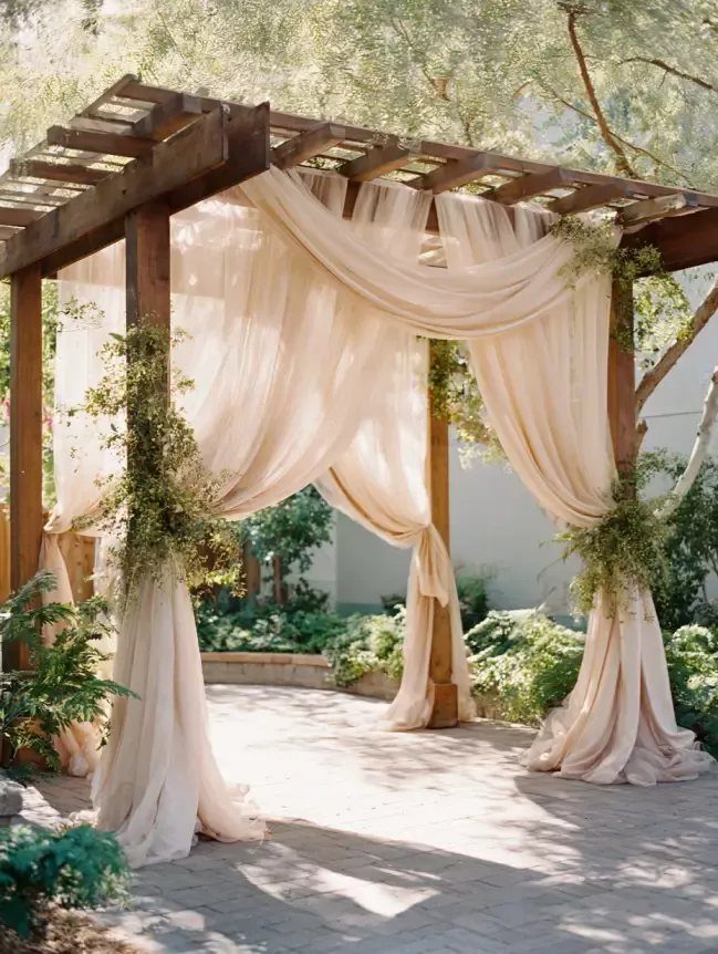 Cream sheer fabric draped over a wooden pergola with green foliage accents outdoors