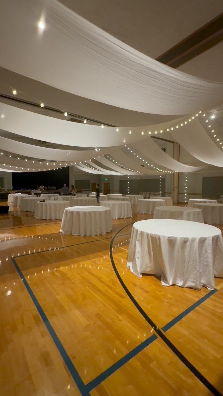 White ceiling drapes with fairy lights over round tables in a gym