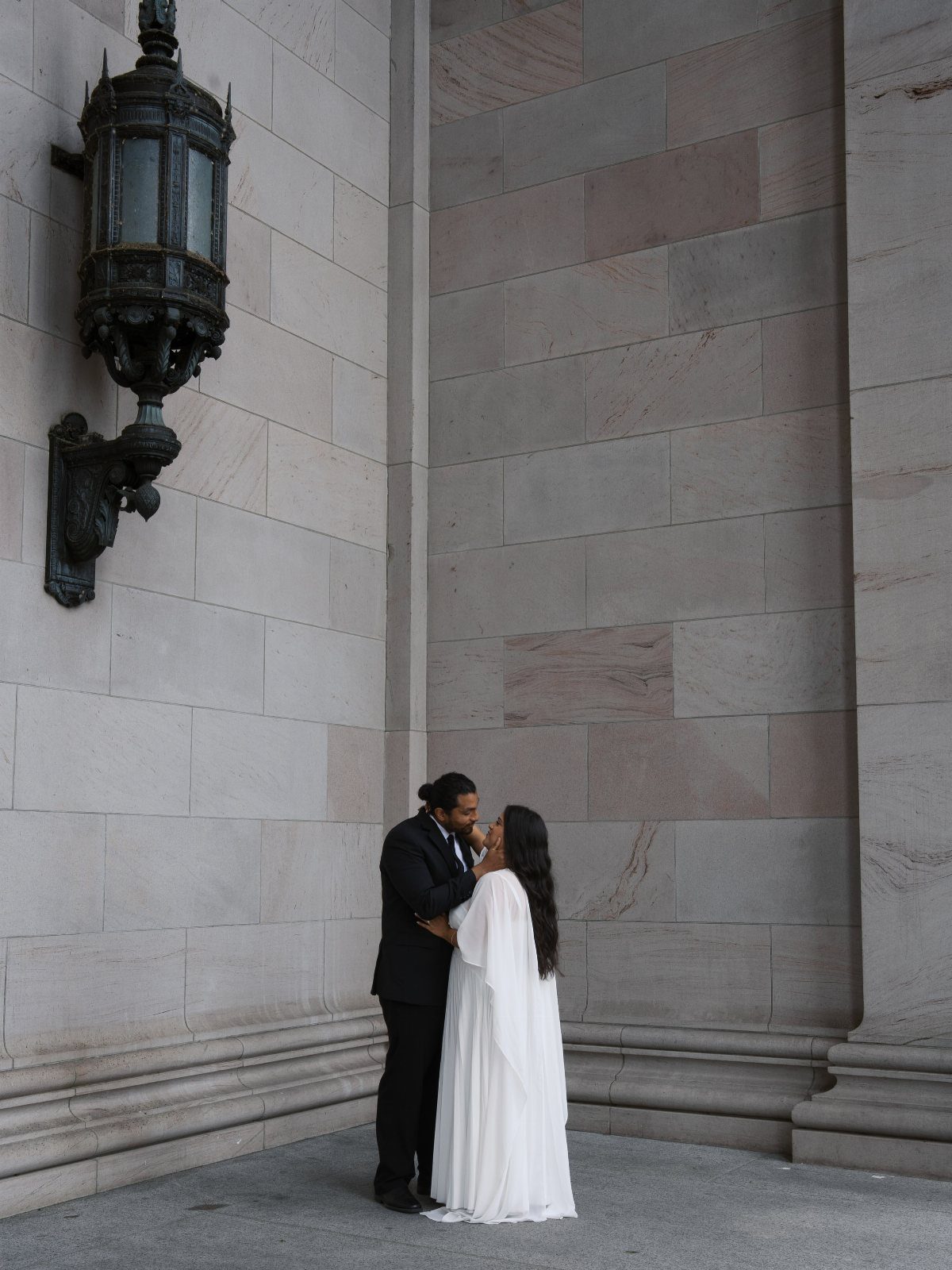 Couple kissing in wedding attire against a grey stone wall with an ornate lantern.