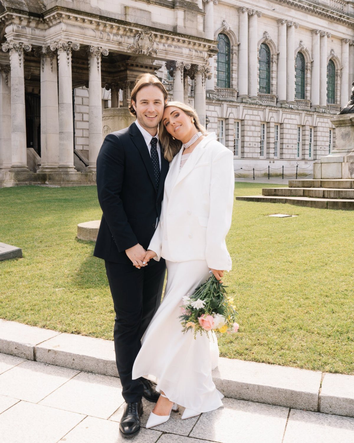 Couple posing in wedding attire, woman wearing a white tailored bridal suit.