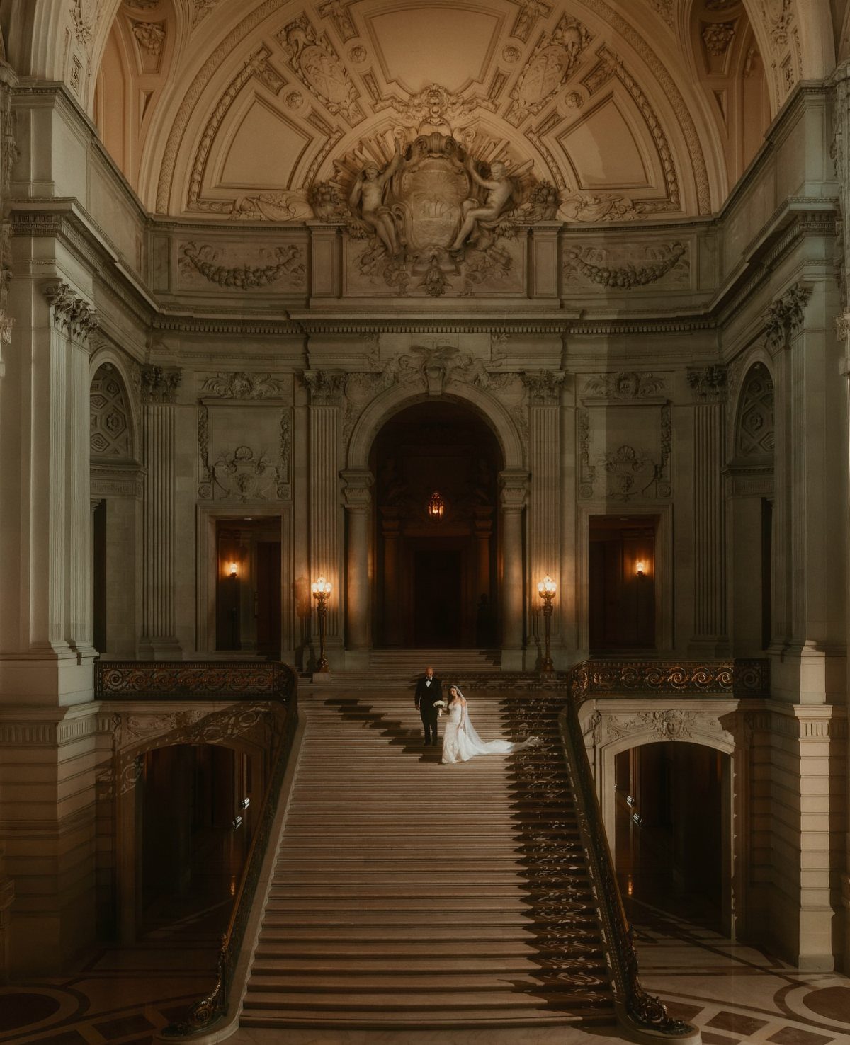 Bride in long white gown and groom on grand ornate stone staircase