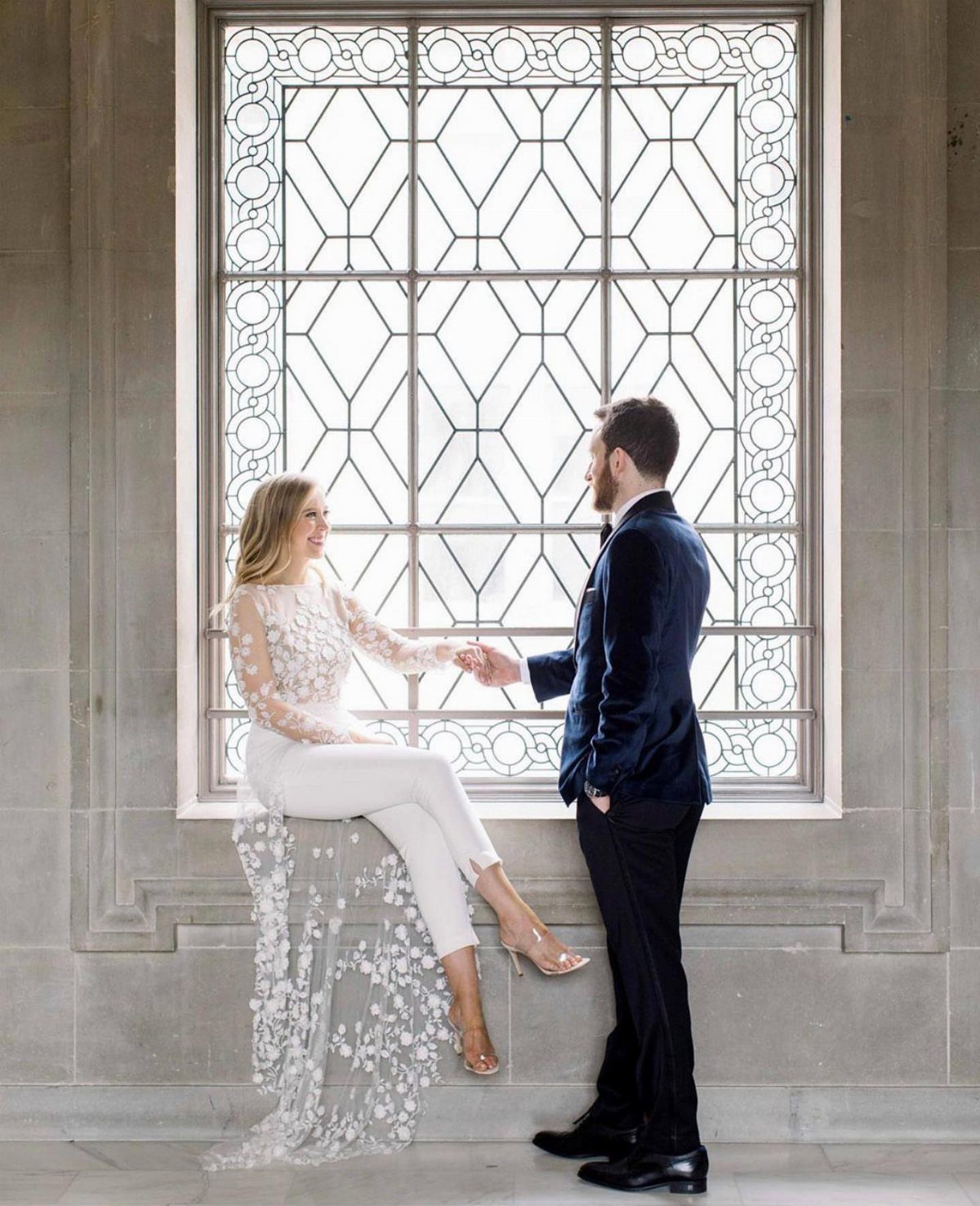 Bride in a white bridal jumpsuit and groom in navy velvet blazer posing together.