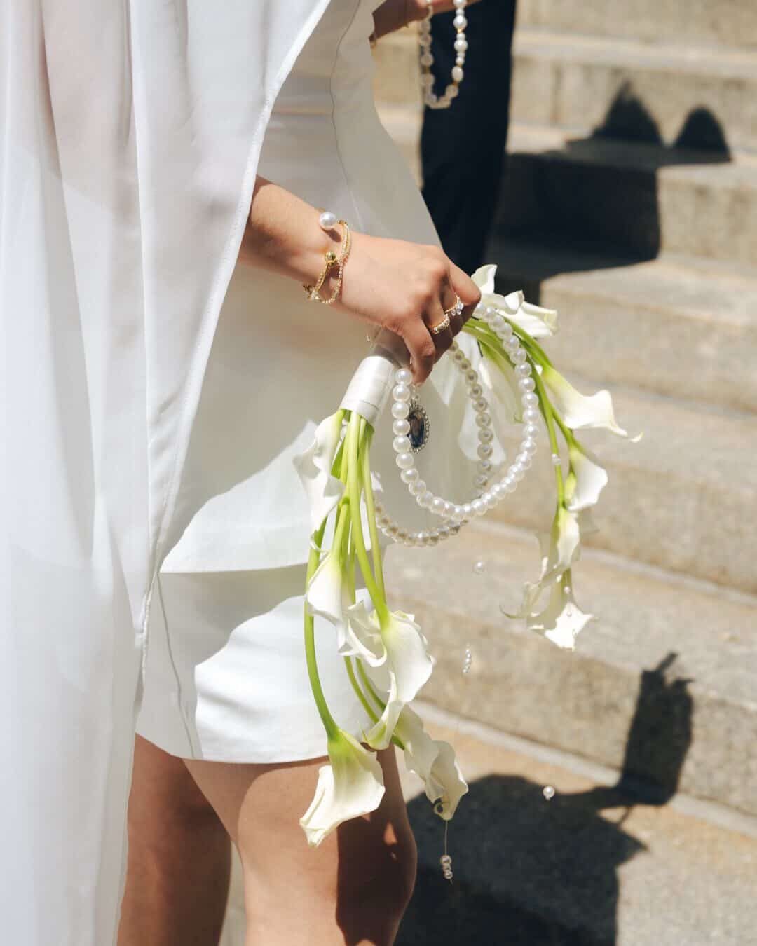 Person in white bridal outfit holding a pearl handbag decorated with calla lilies.