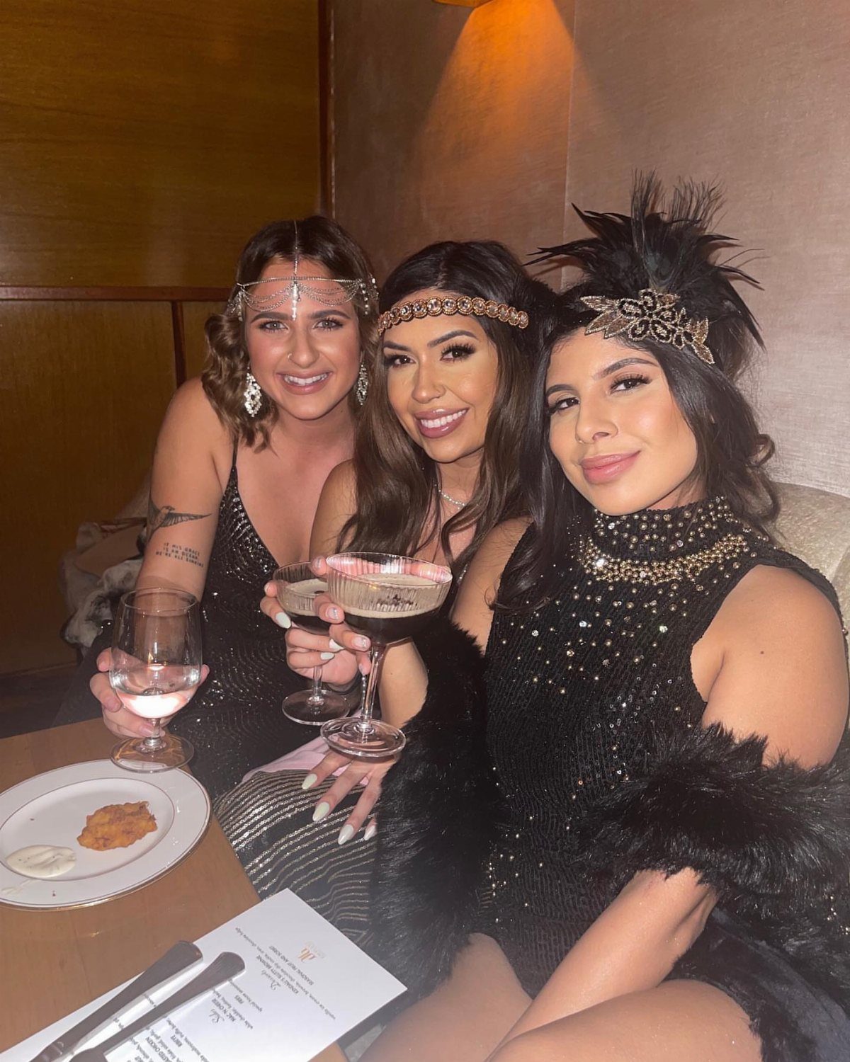 Three women in black sequined flapper dresses and beaded headbands holding cocktail glasses.
