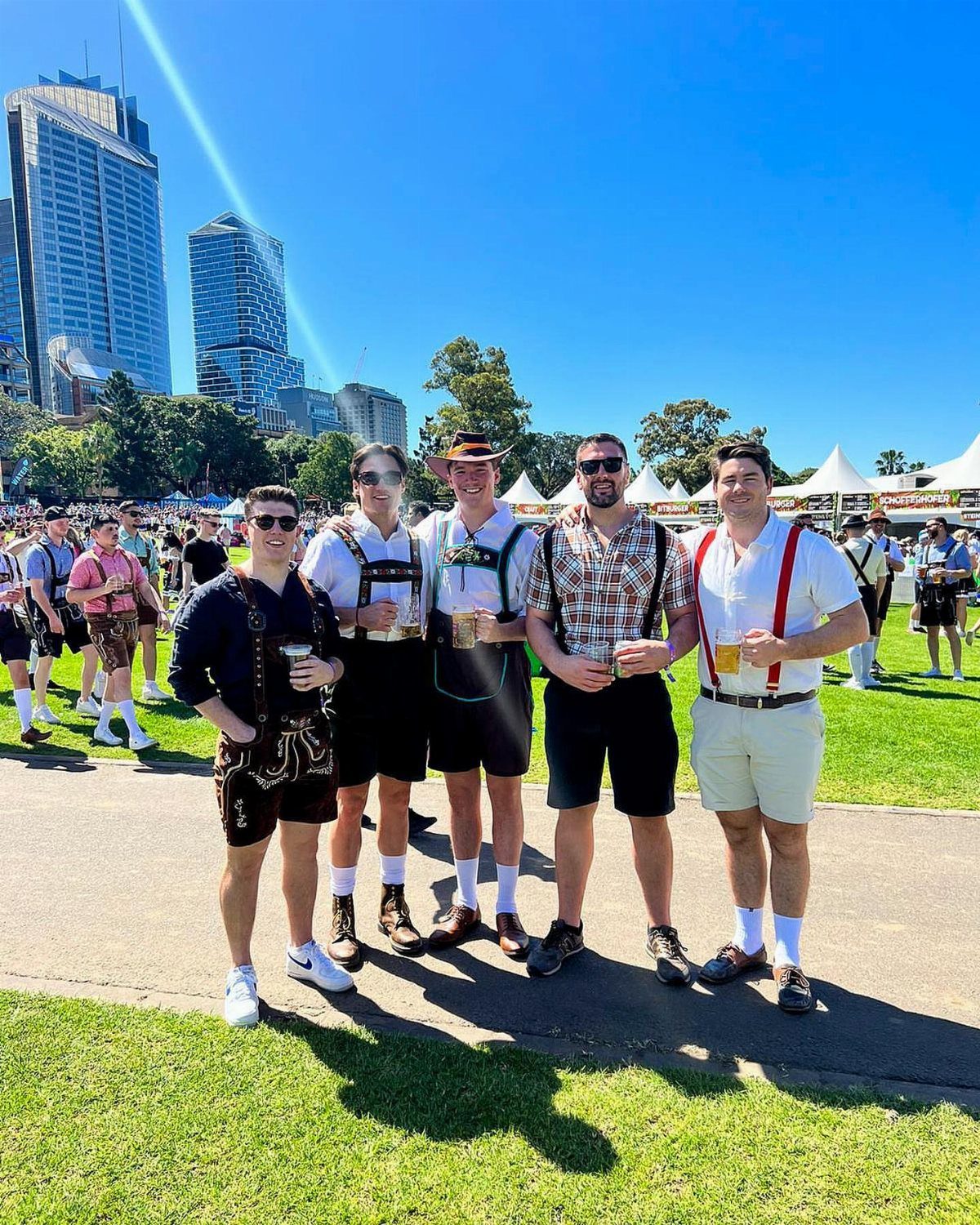Five people wearing traditional German lederhosen and dirndls holding beer mugs outdoors.