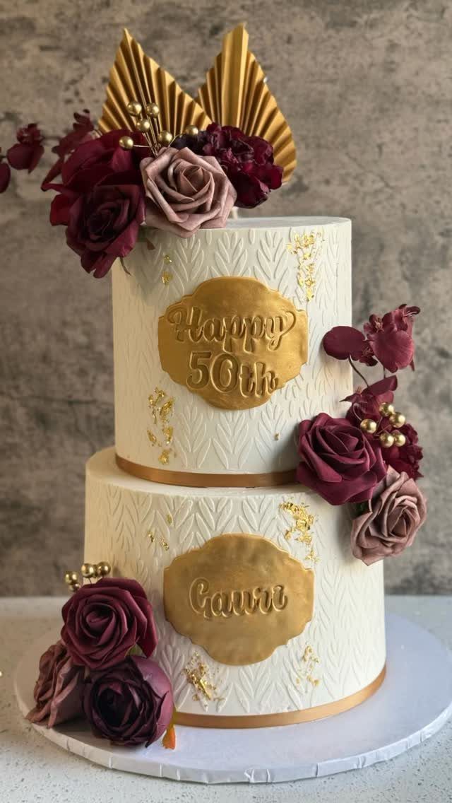 Two-tier white cake with burgundy roses, gold plaques, and textured frosting patterns.