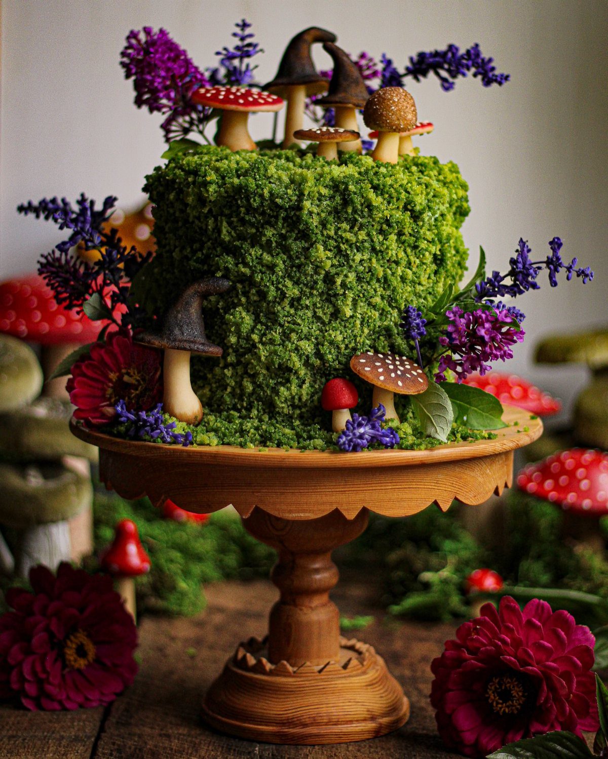 Moss-covered cake decorated with small edible mushrooms and purple flowers on wooden stand