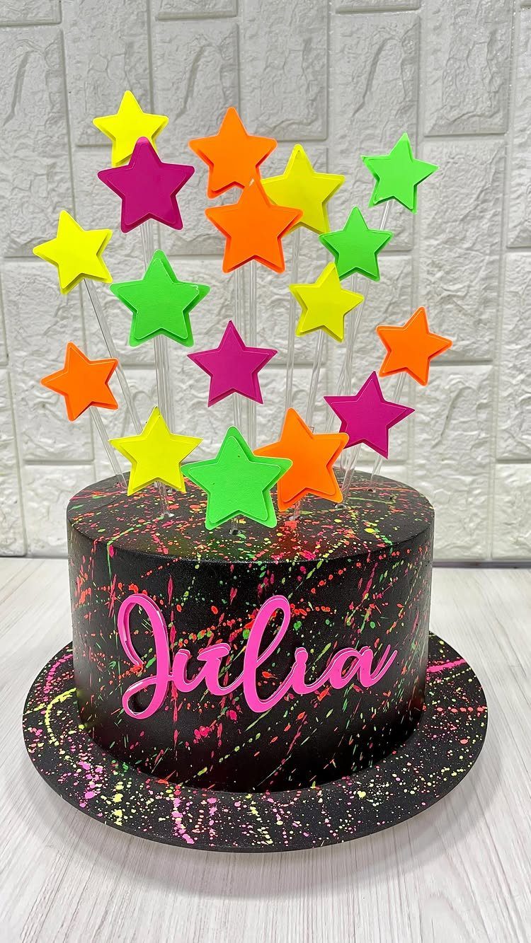 Black cake with neon paint splatters, pink lettering, and colorful star toppers.