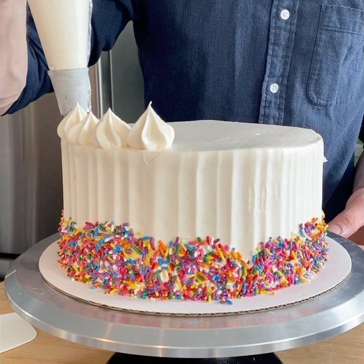 White frosted cake with rainbow sprinkles being decorated with buttercream piping swirls.