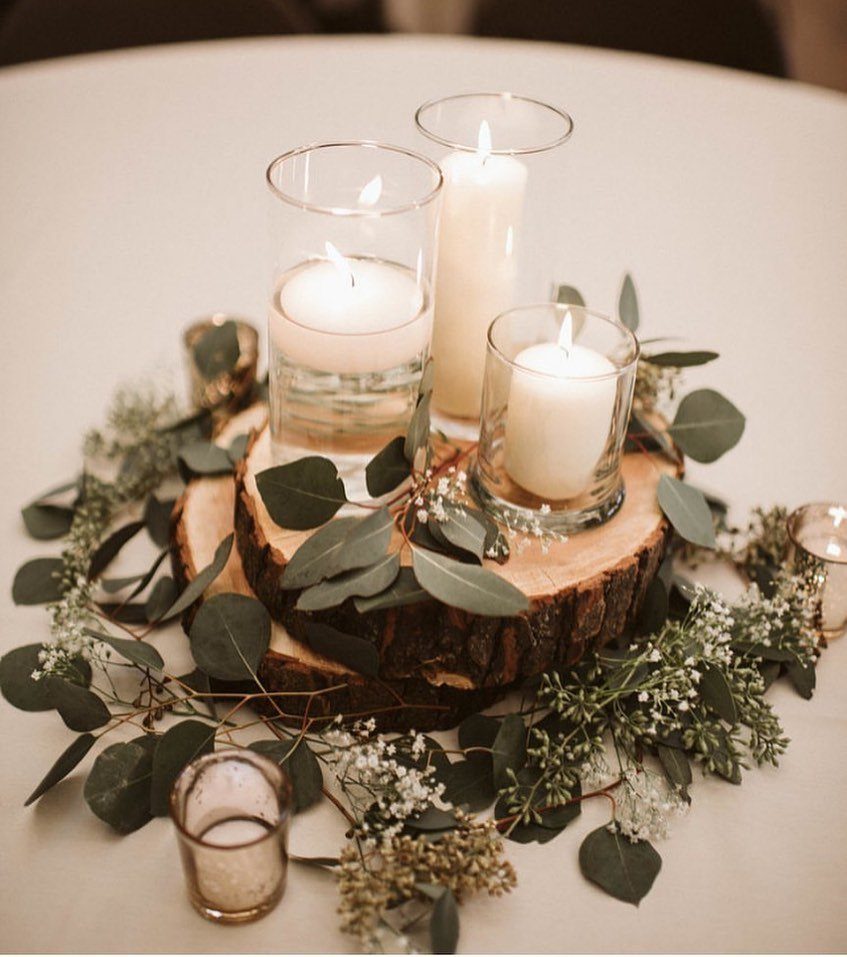 White candles in glass holders on a wood slice with eucalyptus greenery.