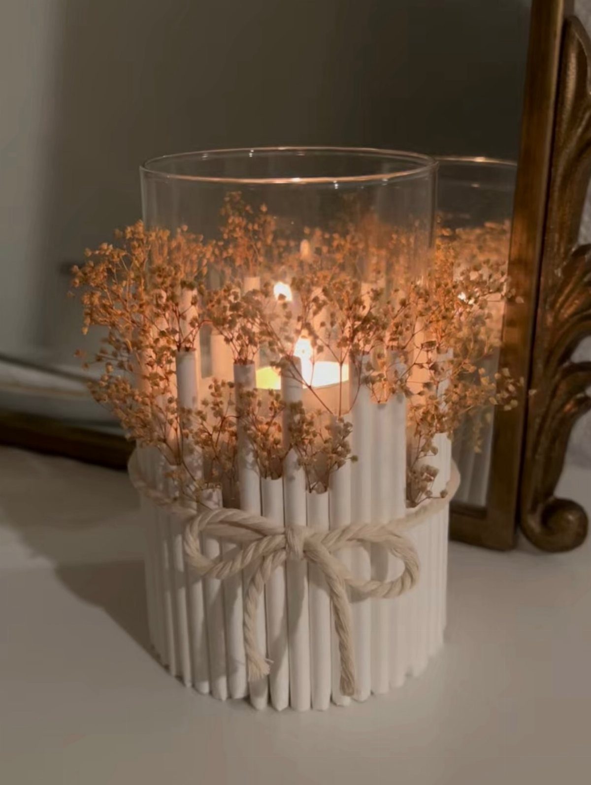 Lit candle in a glass vase wrapped in white sticks and dried flowers
