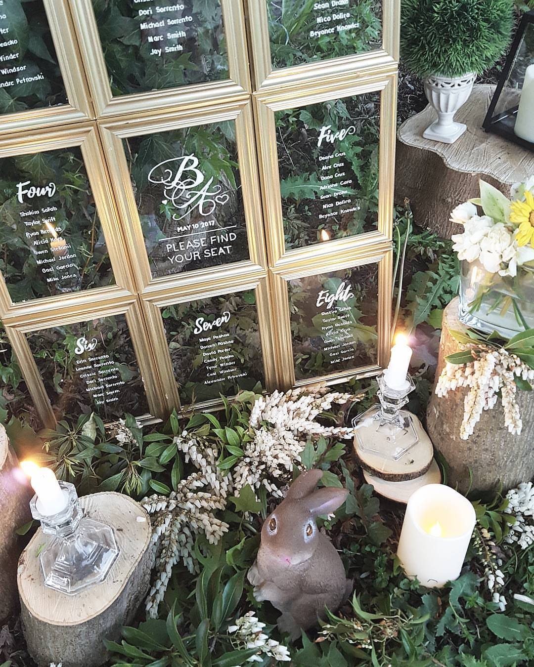 Gold framed seating charts surrounded by greenery, candles on wood slices, and a rabbit.