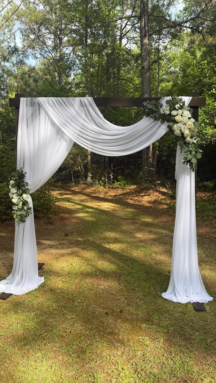 Wooden wedding arch draped in white fabric with white floral arrangements in a forest