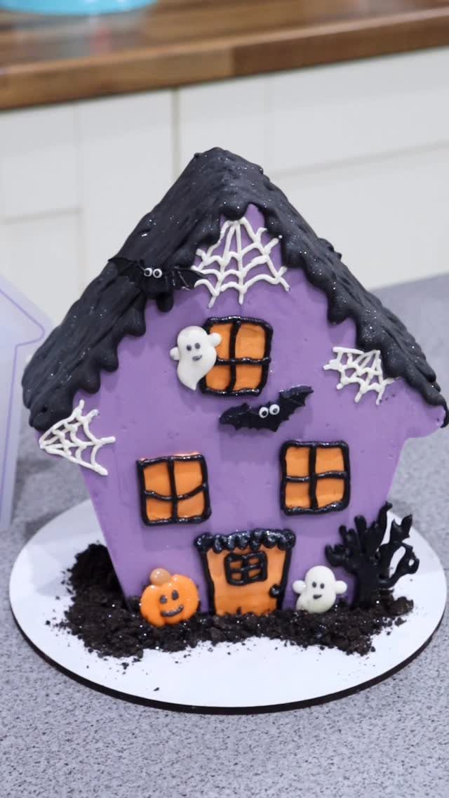 Purple haunted house cake with black roof, ghosts, spiderwebs, and a pumpkin.