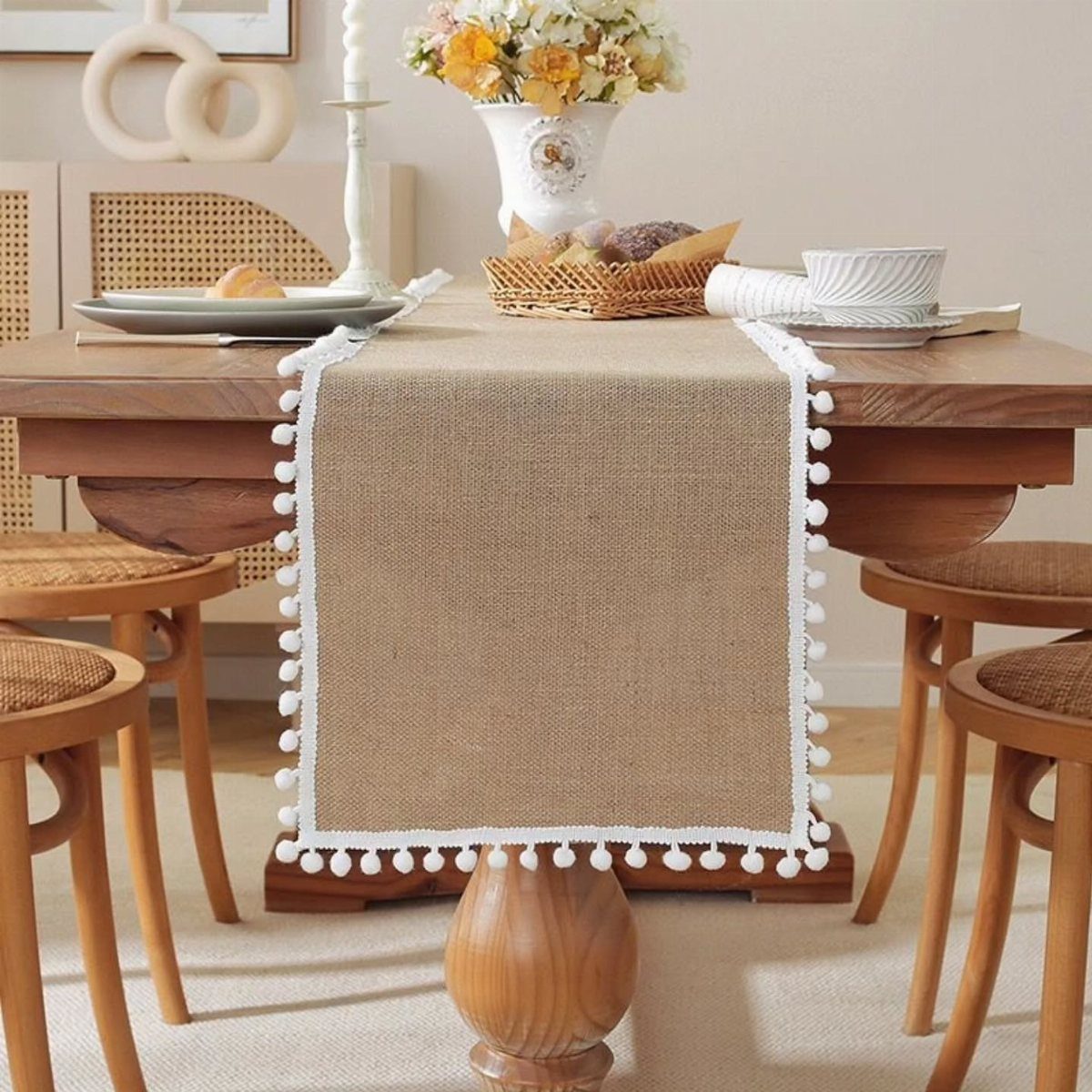 Burlap table runner with white pom-pom trim on a rustic wooden dining table.
