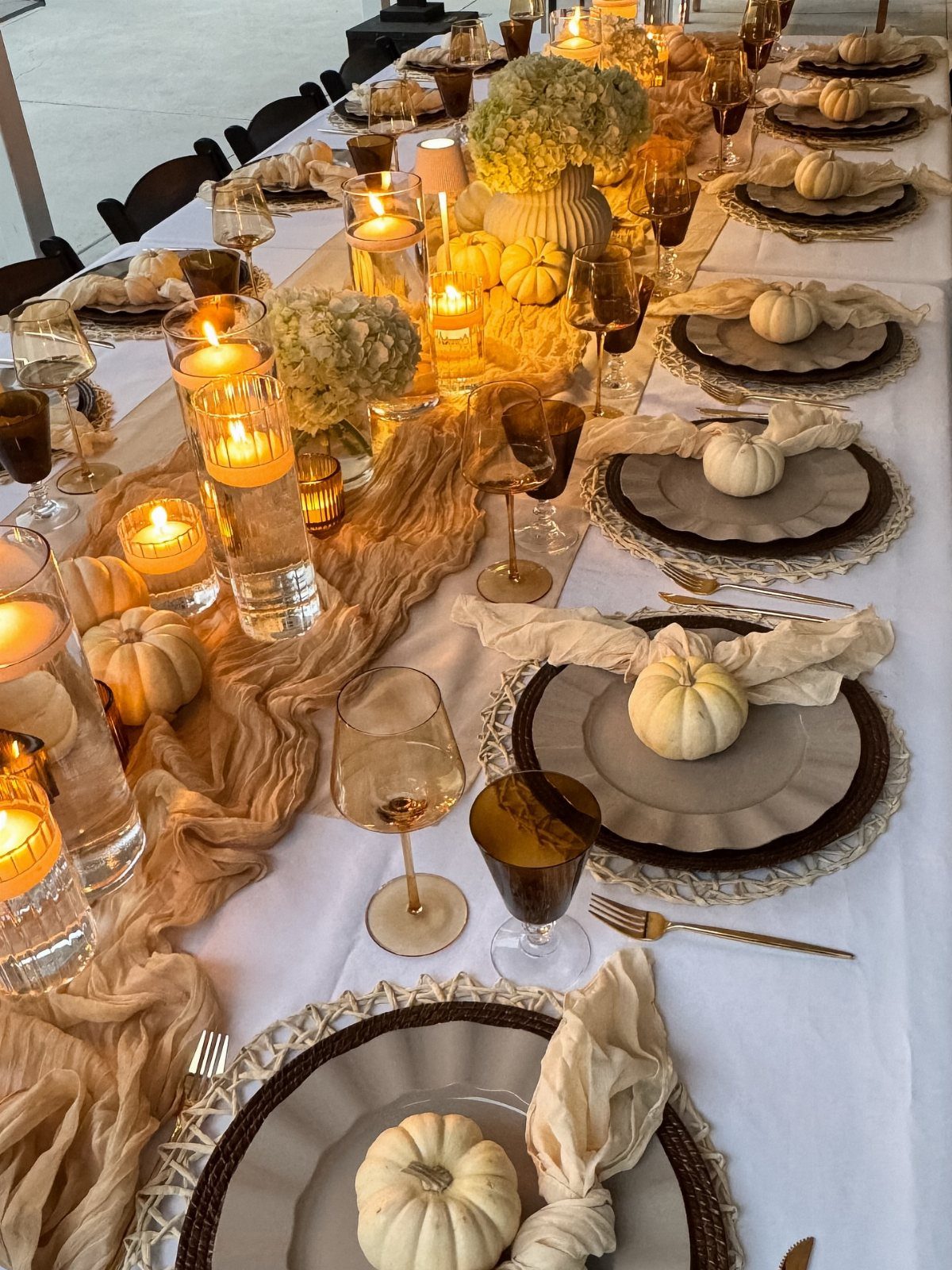 Elegant table setting with white pumpkins, gold cutlery, and glowing candles on white linen.
