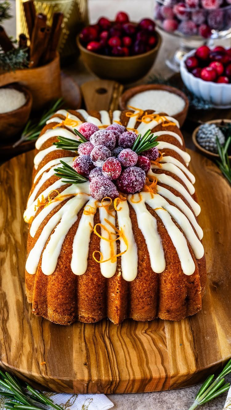 Bundt cake topped with white icing, frosted cranberries, and orange zest on wood.