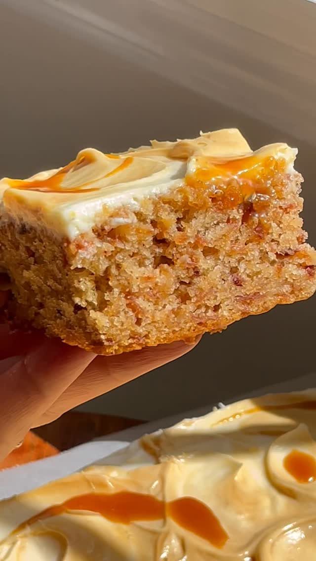 Slice of golden cake with white frosting and caramel drizzle held by hand.