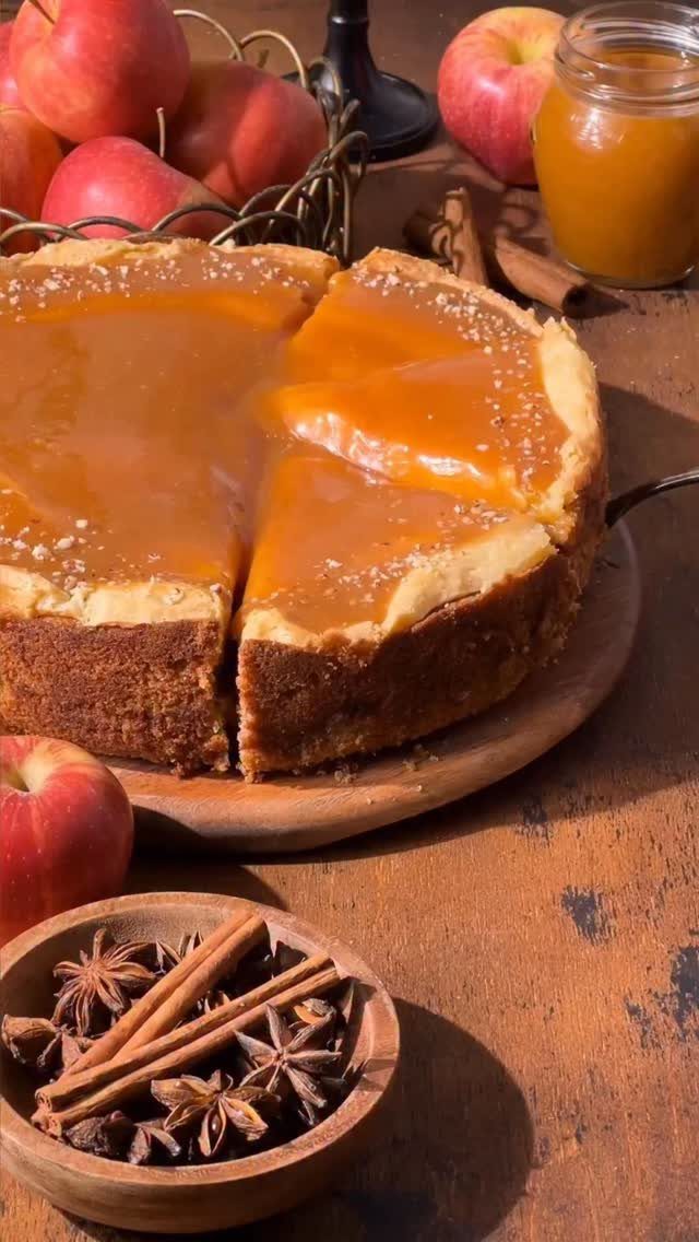 Caramel cheesecake on a wooden board surrounded by red apples and cinnamon sticks.