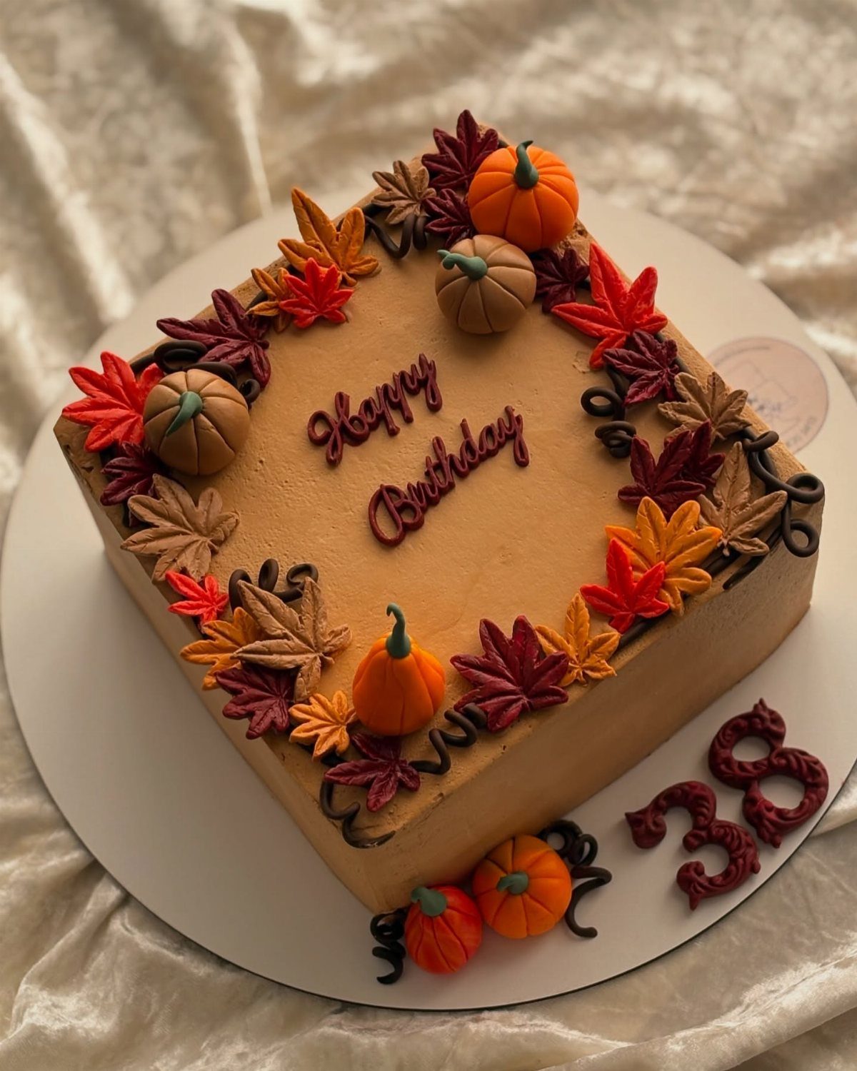Square tan birthday cake decorated with autumn leaves, pumpkins, and chocolate lettering.