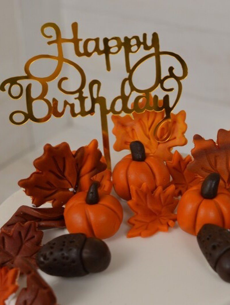 White birthday cake decorated with orange pumpkins, autumn leaves, and gold topper.