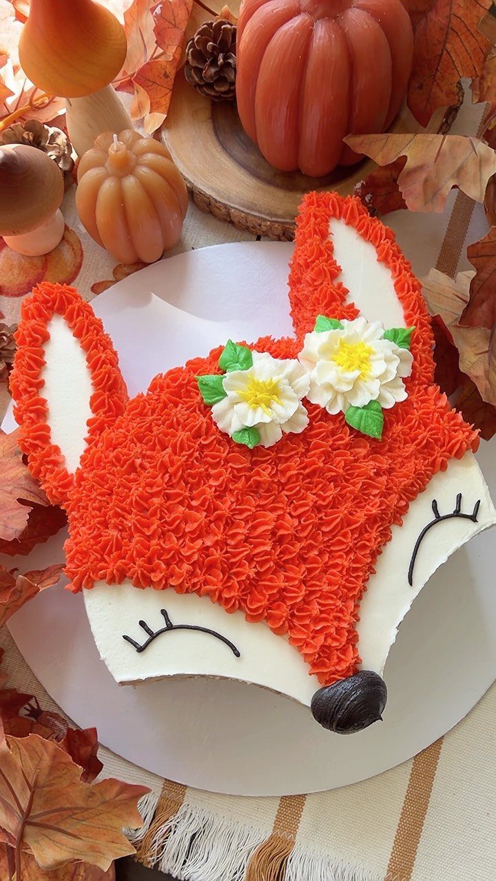 Fox-shaped cake with orange frosting and white flowers surrounded by autumn decorations.