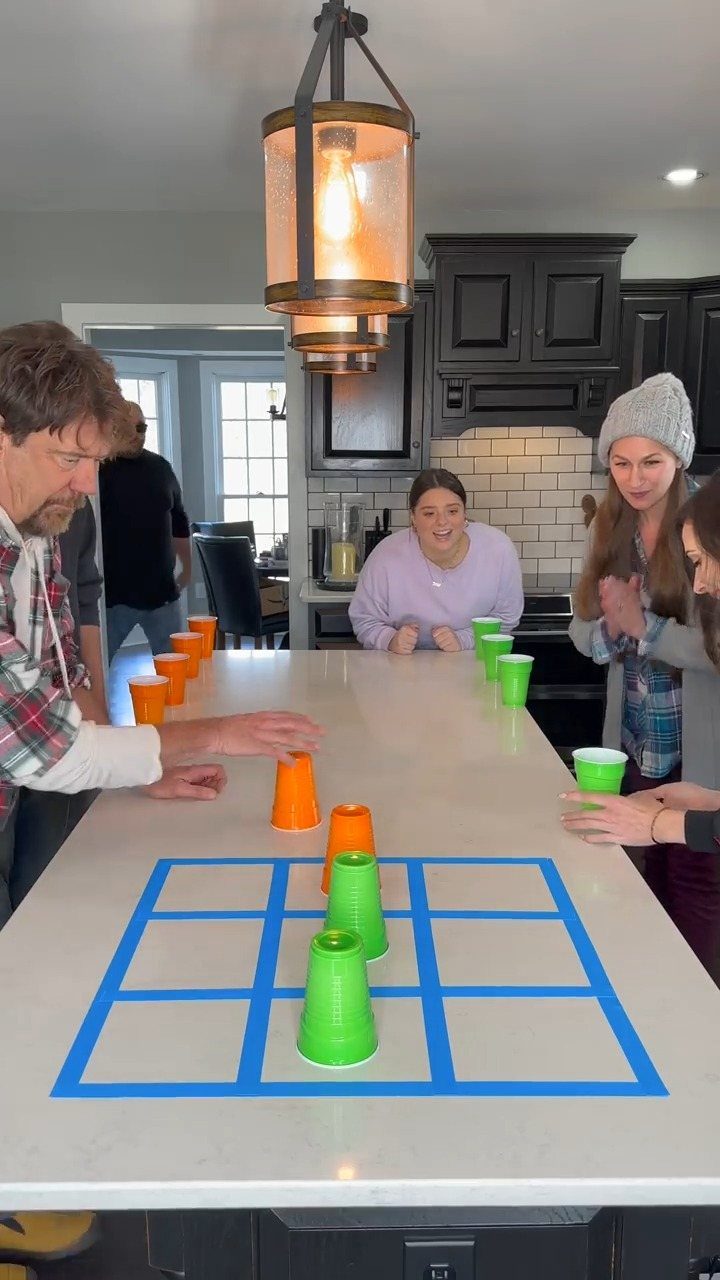 Adults playing a cup game on a kitchen island with a blue tape grid.
