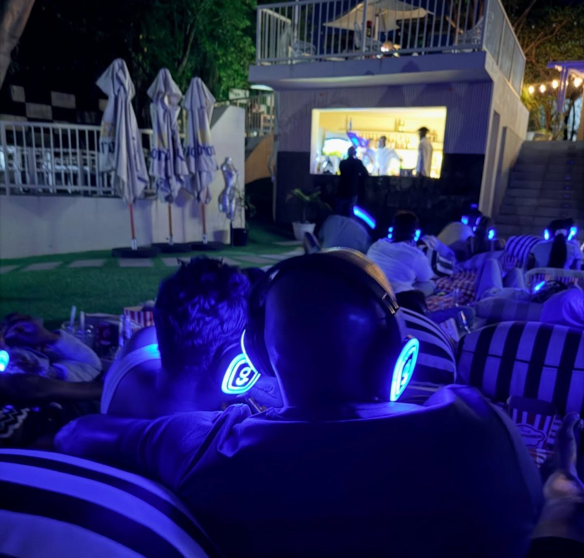 People wearing glowing blue headphones lounging on striped cushions at a night party.