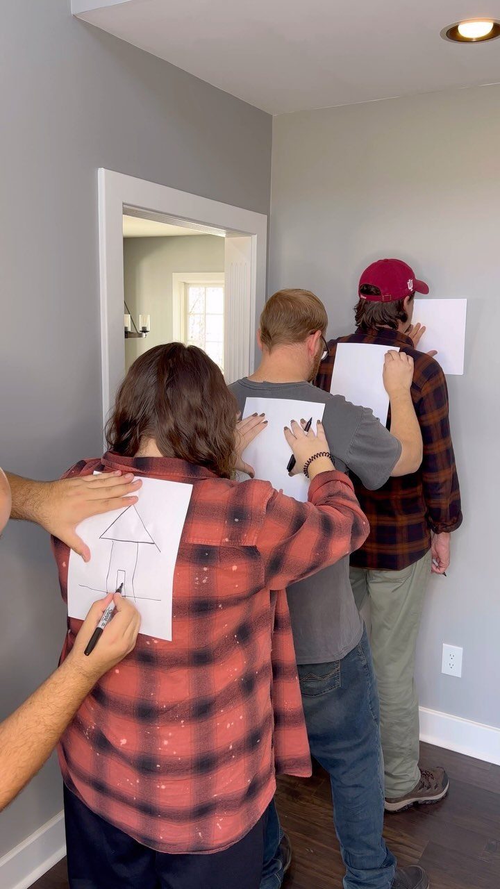People playing a drawing game with paper taped to their backs in a room.