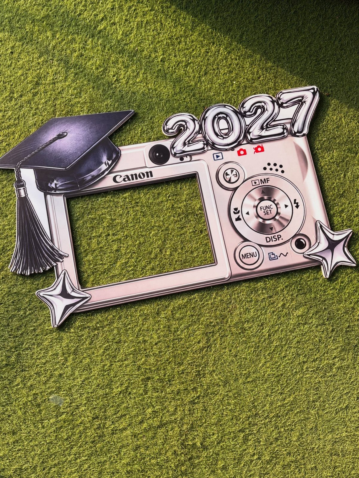 Camera-shaped photo prop frame with graduation cap and 2027 balloons on green grass