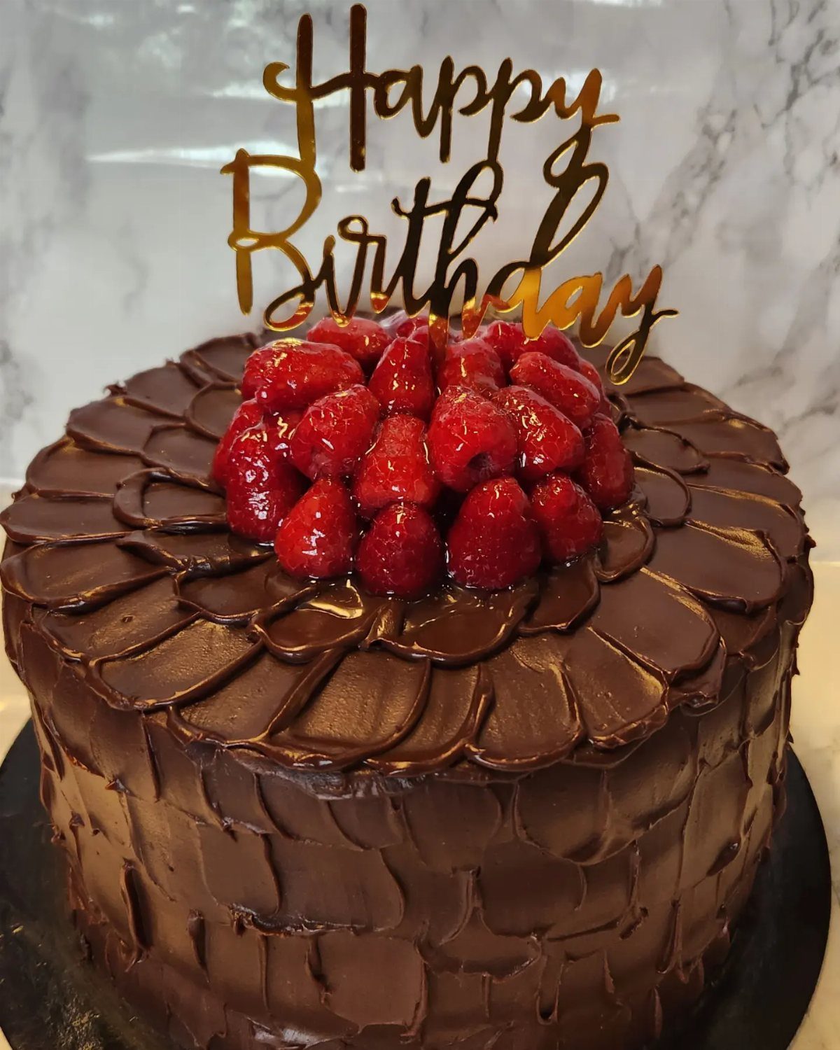 Chocolate birthday cake topped with fresh red raspberries and a gold topper.