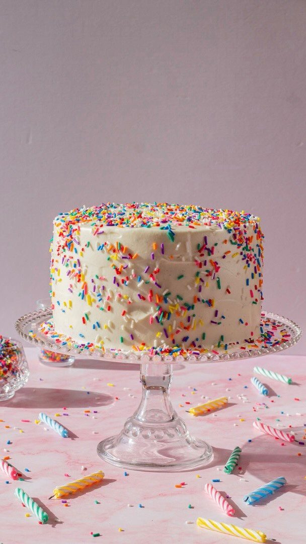 White frosted birthday cake with colorful sprinkles on a glass stand with candles.