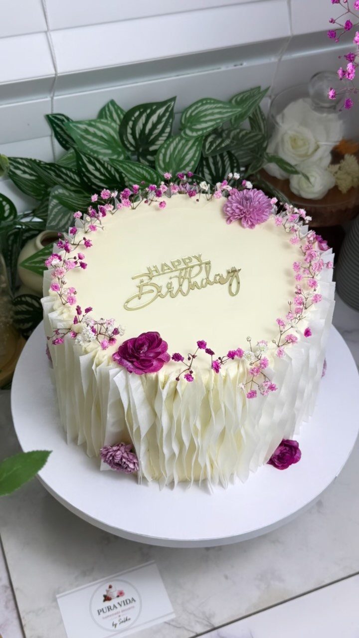 White birthday cake decorated with small pink and purple flowers and gold lettering.