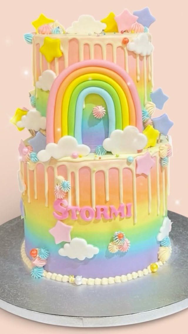 Two-tier rainbow cake with white drips, a 3D rainbow, clouds, and stars.