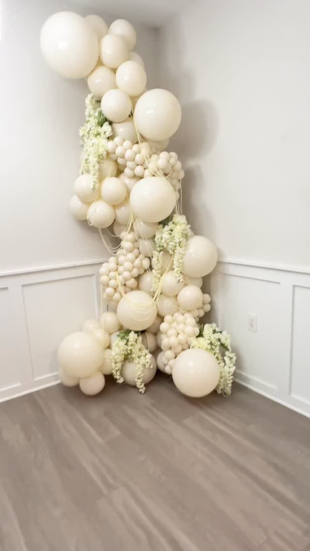 White and cream balloon garland with floral accents in a room corner.