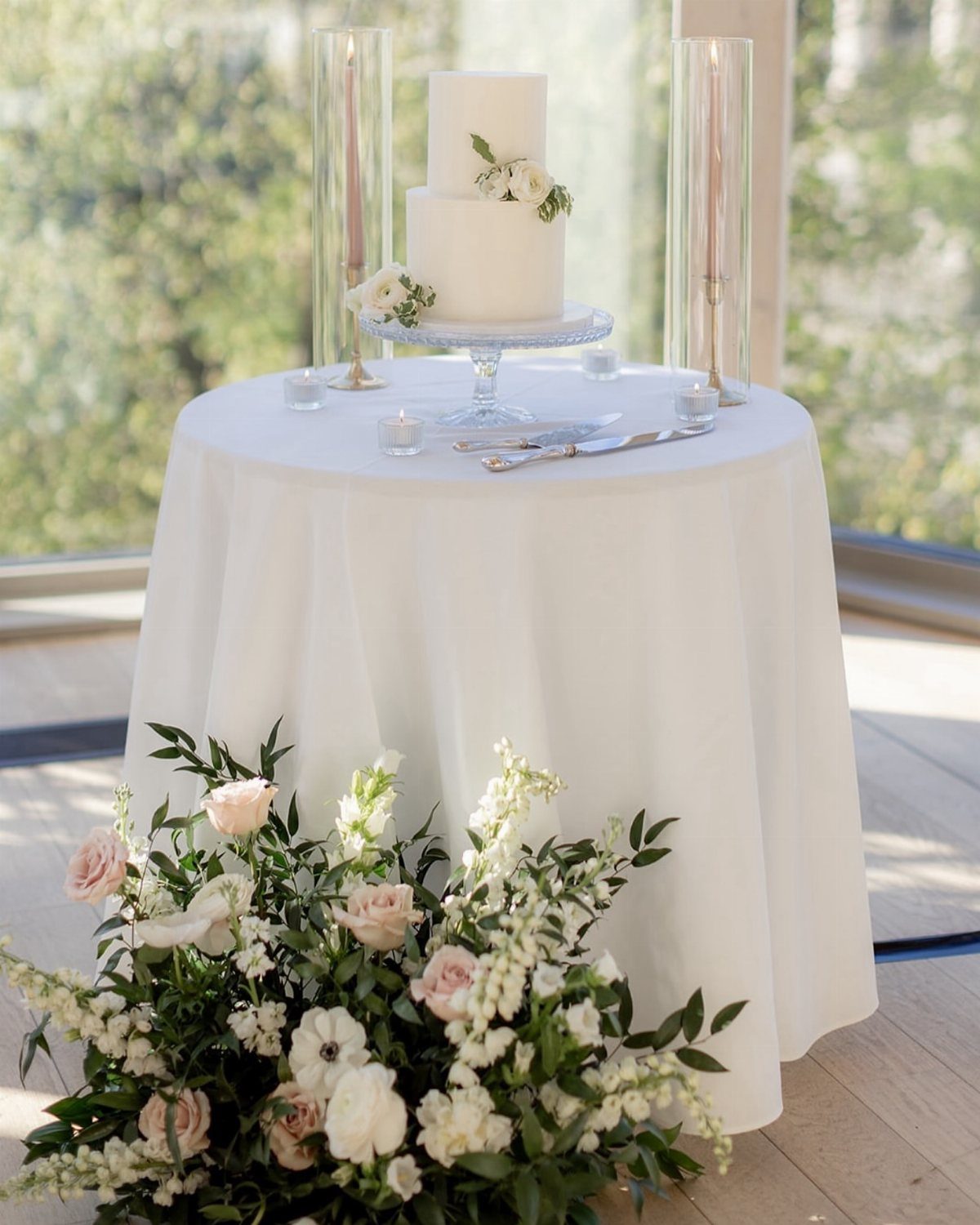 Two-tier white wedding cake on a white draped table with floral arrangements.