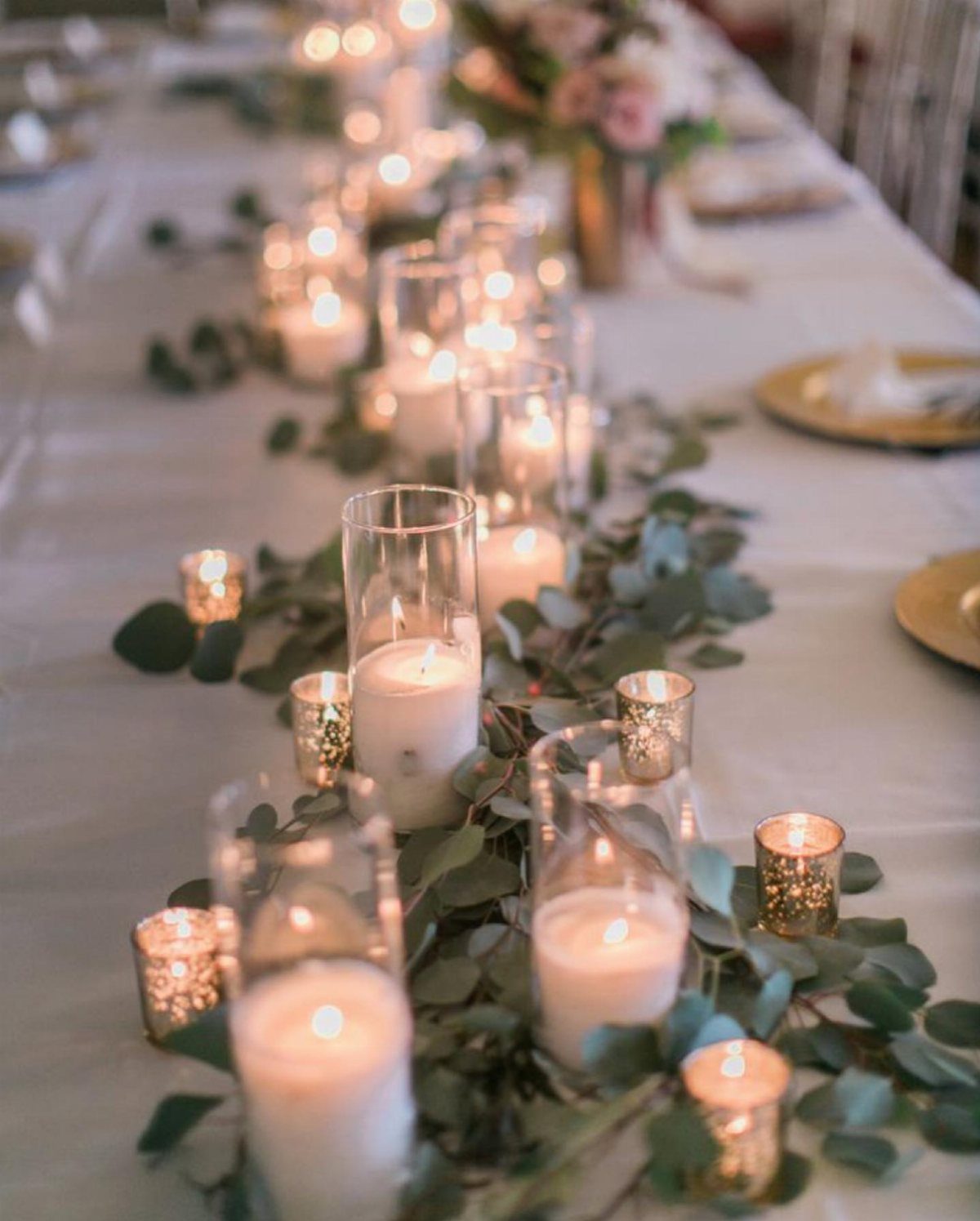 Lit white candles in glass holders and gold votives with eucalyptus leaves on table