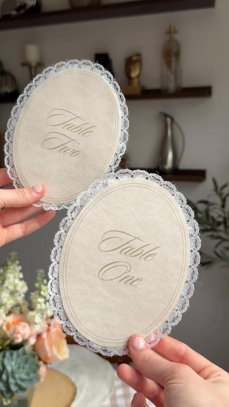 Oval cream table number cards with white lace borders held by two hands.